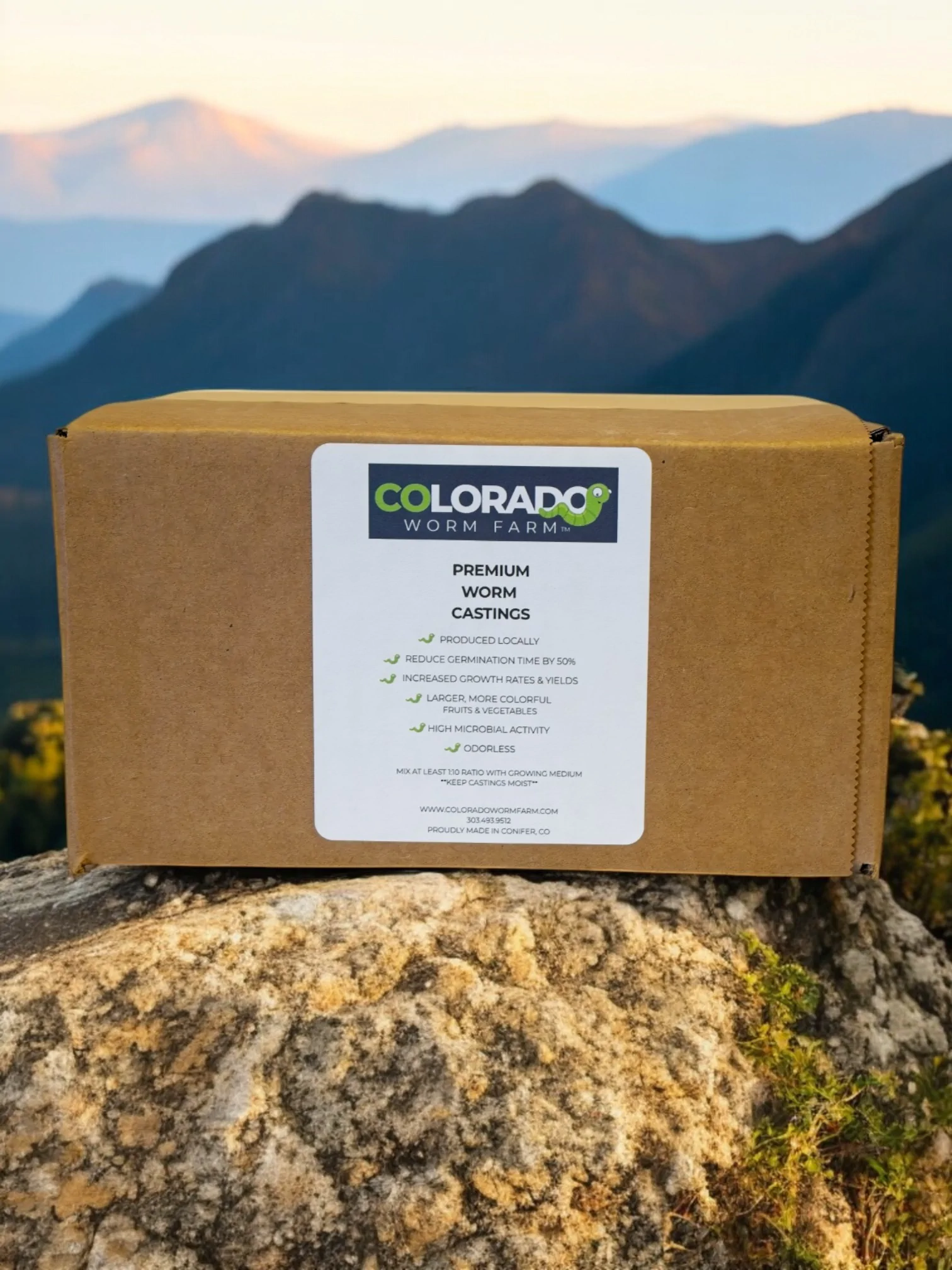 A cardboard box labeled "Colorado Worm Farm Premium Worm Castings" sits on a rock with mountains in the background.