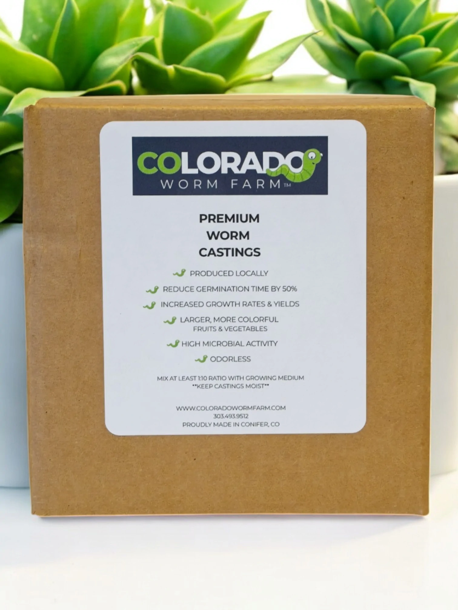 A box labeled "Colorado Worm Farm Premium Worm Castings" stands in front of two green succulent plants, listing product benefits and company details on the front.