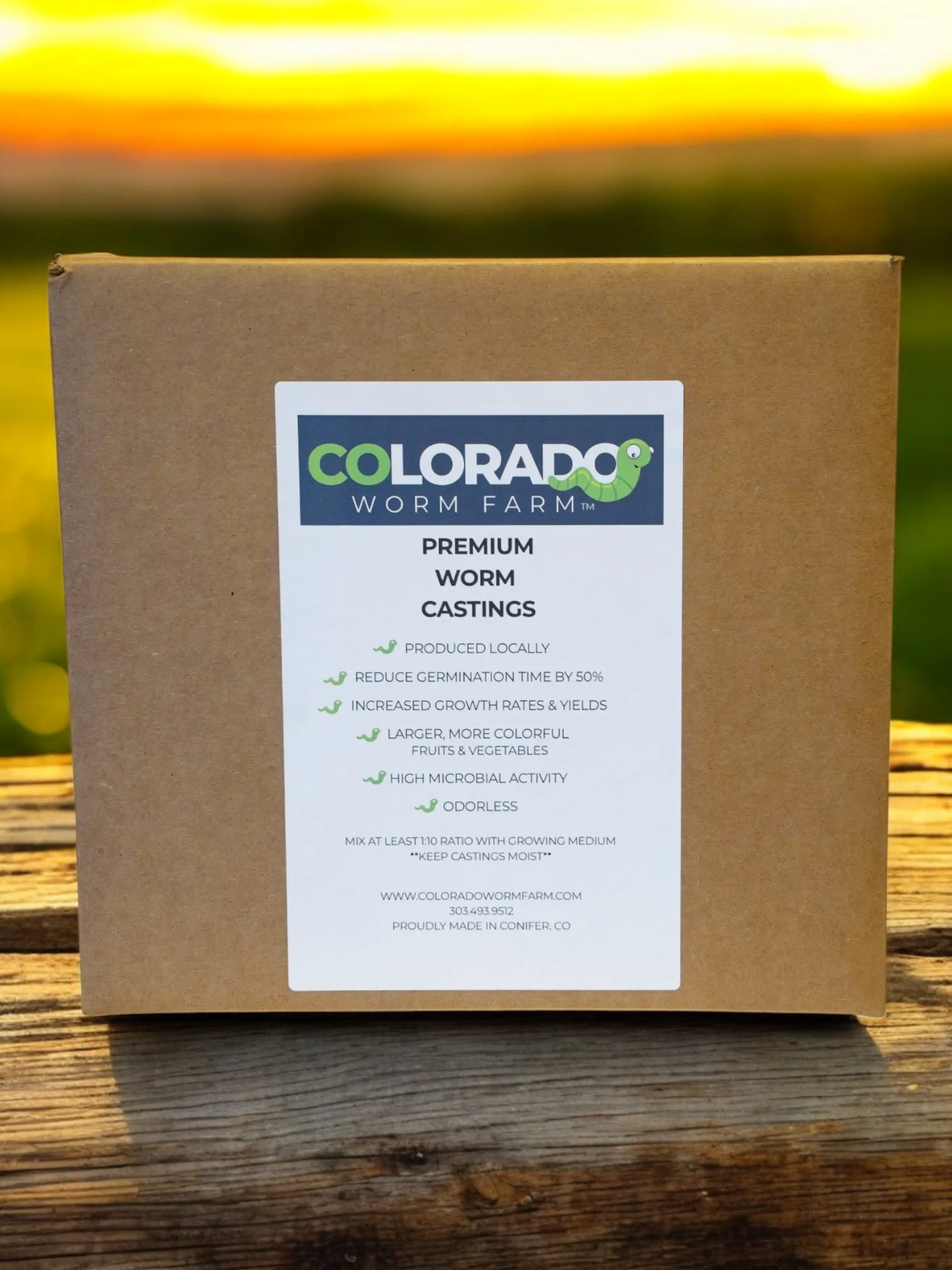 A cardboard box labeled "Colorado Worm Farm Premium Worm Castings" sits on a wooden surface with a blurred outdoor background at sunset.