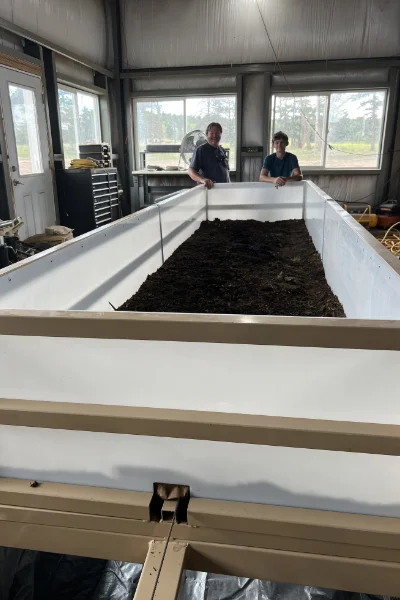 Two people stand on either side of a large rectangular container filled with soil inside a metal industrial building.