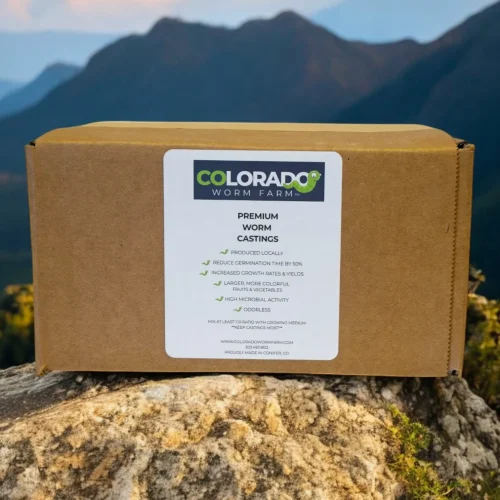 A cardboard box labeled "Colorado Worm Farm Premium Worm Castings" sits on a rock with mountains in the background.