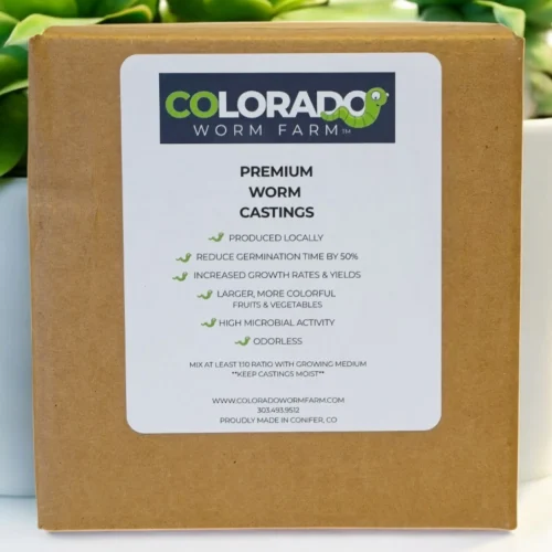 A box labeled "Colorado Worm Farm Premium Worm Castings" stands in front of two green succulent plants, listing product benefits and company details on the front.