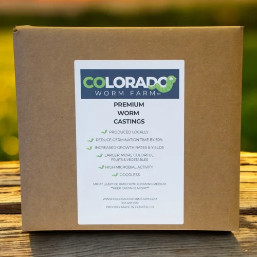 A cardboard box labeled "Colorado Worm Farm Premium Worm Castings" sits on a wooden surface with a blurred outdoor background at sunset.
